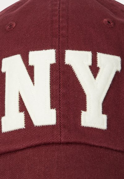 Căciulă de baseball roșie, cu litere albe „NY” ridicate. Fabricată dintr-un material texturat, cu o formă clasică structurată și cu un soare curbat.