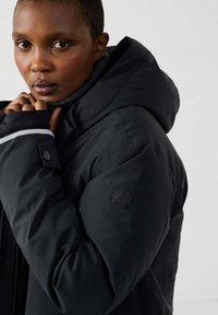 Vrouw met kort haar die een zwarte pufferjack met capuchon draagt, handen bij haar gezicht, recht vooruit kijkend tegen een effen achtergrond.