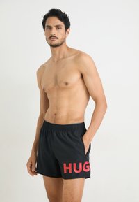 Juodos maudymosi šortai su raudonu "HUG" grafikos dizainu, elastingu juosmeniu, šoninėmis kišenėmis ir lengvu audiniu, tinkamu vandens pramogoms.