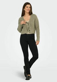 Gilet tricoté vert, coupé, avec un motif en câble et une fermeture à nouer, associé à un jean skinny noir et des mocassins noirs à clous.