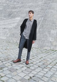 Smešane smeđe kožne cipele sa vezicama, sivi odijela, košulja s uzorkom, crni kaput, stoji na kamenoj površini pored teksturisanog zida.