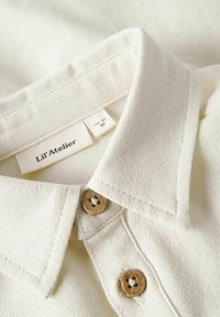 Crèmekleurig shirt met een kraaghals, twee houten knopen en een textielstructuur. Etiket leest "Lil' Atelier."