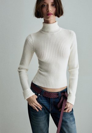 Maglione bianco a collo alto con righe, abbinato a jeans blu scuro con una cintura a strisce bordeaux annodata in vita.