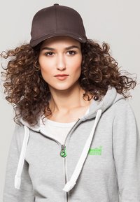 Bruine pet met een gebogen rand, gedragen met een grijze hoodie met ritssluiting, voorzien van witte koorden en een groen logo op de borst.