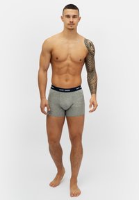 Grå bomullskalsonger med ett marinblått midjeband märkt "HAPPY SHORTS", med en tight passform och sömlös design. Modellen har en tatuering på sin vänstra arm.