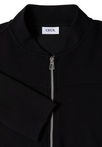 Veste noire zippée avec col montant et fermeture éclair argentée, avec une étiquette blanche portant le nom de la marque "CECIL" à l'encolure.