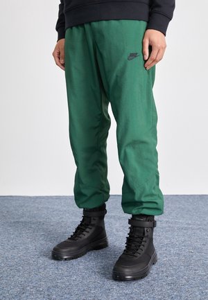 Person trägt grüne Nike-Trainingshose und schwarze Schnürstiefeletten und steht auf einem blau carpetten Boden.