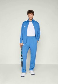 Trainingspak in helderblauw polyester met een ritsjack en broek met elastische tailleband. Witte t-shirt eronder, met een tennisracket in de hand.