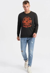Felpa nera con grafica arancione "Chicago State", maniche lunghe, polsini a coste, abbinata a jeans chiari strappati e sneakers bianche.