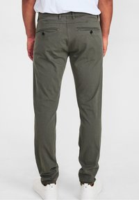 Man som bär grå skräddarsydda byxor och vita sneakers står mot en neutral bakgrund, bakifrån synlig.