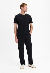 Zwarte gestructureerde t-shirt met korte mouwen en ronde hals, gecombineerd met zwarte broek en witte sneakers; model staat casual met één hand in de zak.