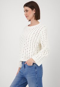White knitted sweater featuring an open lattice design, round neckline, and long sleeves, paired with blue denim jeans.