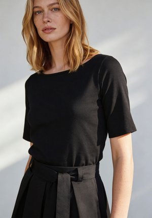 Frau mit schulterlangen blonden Haaren, die ein eng anliegendes schwarzes Kurzarmoberteil und eine hoch taillierte schwarze Hose mit Stoffgürtel trägt.