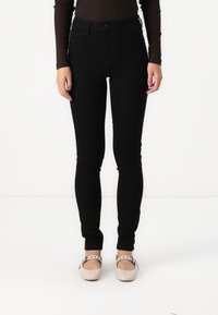 Μαύρα skinny τζιν με ψηλή μέση, κατασκευασμένα από τζιν, με κλείσιμο με κουμπί και μπροστινές τσέπες, συνδυασμένα με ανοιχτόχρωμα παπούτσια.