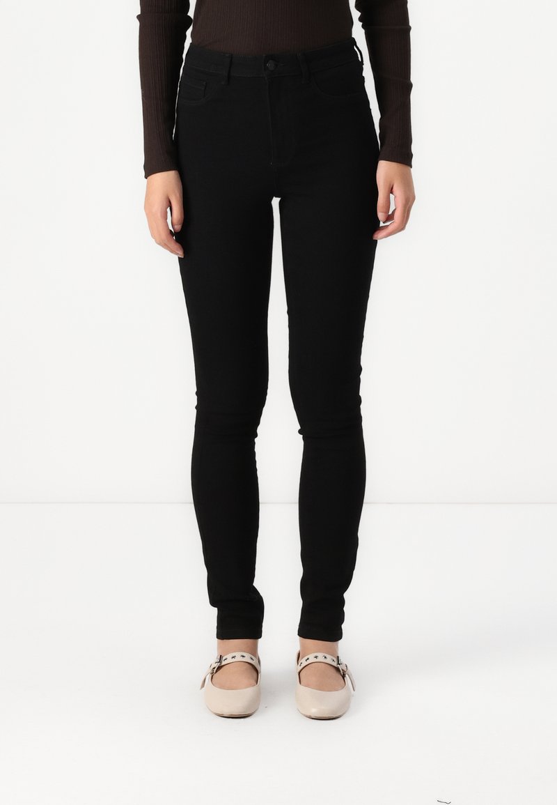 Μαύρα skinny τζιν με ψηλή μέση, κατασκευασμένα από τζιν, με κλείσιμο με κουμπί και μπροστινές τσέπες, συνδυασμένα με ανοιχτόχρωμα παπούτσια.