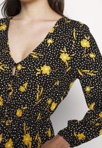Vestido negro con lunares blancos y estampado floral amarillo, con un escote en V profundo, cierre de botones y mangas largas abullonadas. Tejido suave.