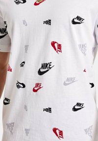 T-shirt in cotone bianco, caratterizzata da una stampa ripetuta di loghi Nike in nero, rosso e grigio e dal testo "Just Do It" in diverse dimensioni.