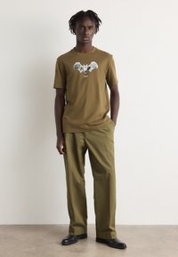 Olive green t-shirt with bat graphic and "BOSS" text, paired with olive green pants. Model stands on a neutral background. Black shoes.