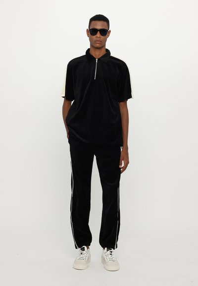 Schwarzes Velours-Zip-Up-Polo mit kurzen Ärmeln, kombiniert mit schwarzen eng geschnittenen Hosen mit weißen Seitestreifen und weißen Sneakers.