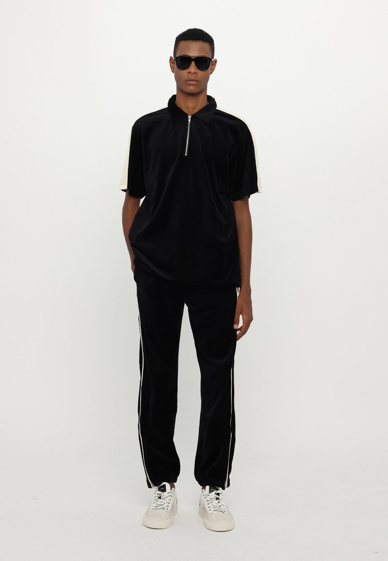Polo noir en velours zippé à manches courtes, associé à un pantalon noir fuselé avec des rayures blanches sur les côtés et des baskets blanches.