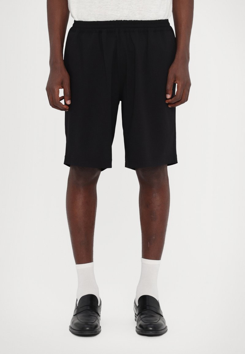 Shorts neri con elastico in vita, lunghi fino al ginocchio, realizzati in un tessuto liscio. Abbinati a calzini bianchi e scarpe nere senza lacci.