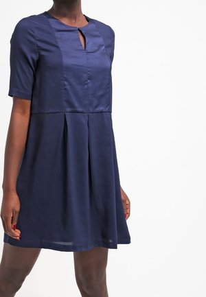 Kvinde iført en mørkeblå, knælange kjole med korte ærmer og en nøglehulshalsudskæring, stående foran en ensfarvet hvid baggrund.