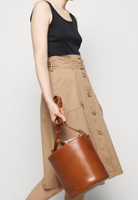 Tan A-lijn rok met knoopdetails en riem, gecombineerd met een zwarte tanktop; bruine lederen emmer tas in de rechterhand gehouden.