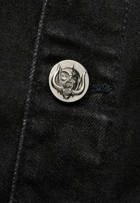 Svart tyg med en texturerad yta och en silverfärgad metallknapp med ett dödskallemönster och böjda horn.