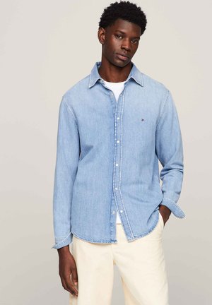 Camicia di denim azzurro chiaro con bottoni, colletto, maniche lunghe, con un piccolo logo sul lato sinistro del petto. Indossata sopra una maglietta bianca.