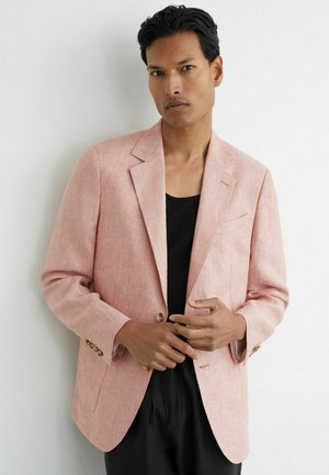 Man met zwart haar die een lichtroze blazer draagt over een zwart overhemd, staand tegen een witte muur, de knopen van de blazer verstelend.