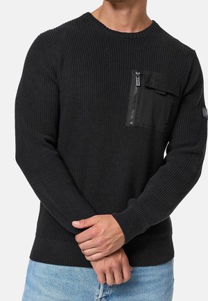 Man draagt zwarte geribde gebreide trui met ritssluiting en klepzak op de borst en lichtblauwe spijkerbroek, handen voor zich geklemd.