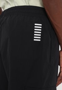 Gros plan d'un pantalon noir avec une ceinture élastique, montrant sept bandes blanches horizontales sur le côté près de la main d'une personne.