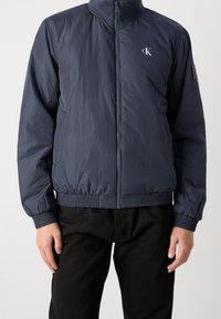 Granatowa kurtka bomberka z suwakiem z przodu, elastycznymi mankietami i paskiem oraz małym logo na klatce piersiowej. Wykonana z lekkiego materiału.