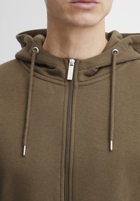 Brun zip-up hette med foret hette, sølvglidelås og matchende snorer. Myk tekstur og en tettsittende design.
