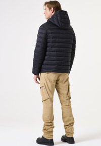 Zwarte pufferjack met horizontale stiksels en een capuchon, gecombineerd met tan cargobroek met zijzakken en zwarte laarzen.