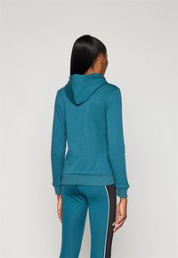 Felpa teal con cappuccio regolabile, polsini e orlo a coste, indossata con leggings coordinati con strisce laterali. Tessuto morbido, vestibilità casual.