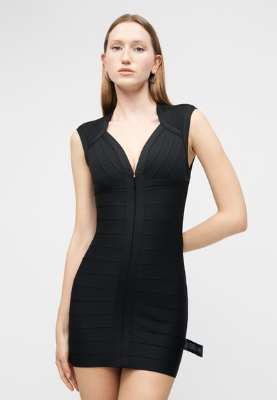 Młoda kobieta o prostych blond włosach, ubrana w dopasowaną czarną sukienkę bez rękawów z zamkiem z przodu i kolczykami kołami, na neutralnym tle.