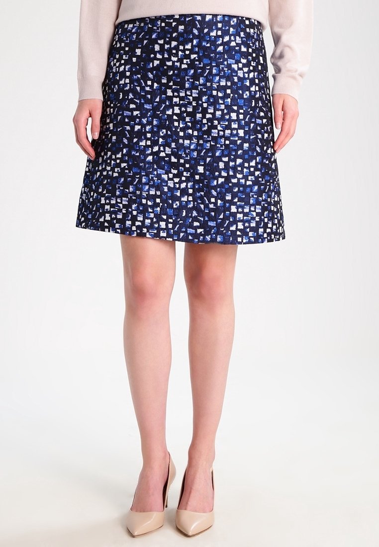 Jupe trapèze longueur genou avec motif abstrait de carrés bleus, noirs et blancs, portée avec un haut beige et des talons pointus couleur nude.