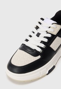 Zapatillas negras y crema que presentan una parte superior de cuero texturizado y textil de patrón, cordones blancos y sutiles acentos crema en la suela.