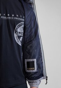 Marineblaue Langarmbluse mit Grafikdruck, ausgestattet mit Ärmeln aus Netzstoff und einem Logo-Patch am linken Arm. Mit elastischen Bündchen und Reißverschlussdetails.