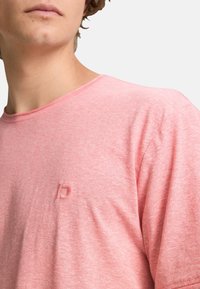 Camiseta de manga corta y cuello redondo en suave tejido rosa con una sutil textura jaspeada y un logo grabado de 'D' en el pecho.