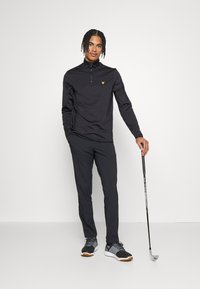 Zwarte rits-top met lange mouwen en een geel logo, gecombineerd met donkere broek en zwarte schoenen. Houdt een golfclub vast, staande tegen een witte achtergrond.