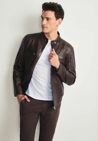 Mężczyzna w ciemno-brązowej skórzanej kurtce na białej koszuli i ciemnych spodniach, stojący przy jasnej ścianie, patrzący w bok.
