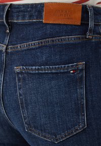 Mørkeblå denimjeans med et skinnmerke fra Tommy Hilfiger, med oransje søm og et lite rødt, hvitt og blått logo på lommen.