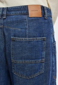 Σκούρο μπλε τζιν με πίσω τσέπη και καφέ δερμάτινη ετικέτα που γράφει "MOS MOSH". Διαθέτει πορτοκαλί ραφές και σχέδιο με ευθεία κοπή.