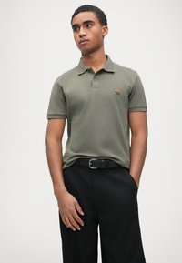 Polo vert olive avec patte de boutonnage à deux boutons, manches courtes et logo brodé marron. Porté avec un pantalon noir et une ceinture noire.