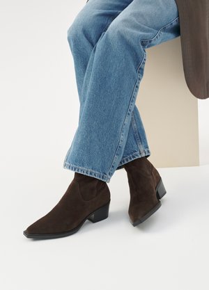 Legs crossed wearing light blue jeans and dark brown suede ankle boots with low block heels against a plain background.