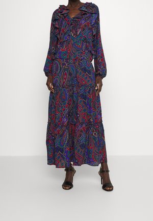 Femme portant une robe à manches longues et longueur cheville avec des motifs cachemire bleus, rouges et violets, et des talons noirs ouverts à l'avant.