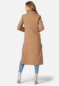 Cappotto lungo in cammello con tessuto testurizzato, vestibilità su misura, colletto a revers e tasche laterali, abbinato a un jeans chiaro e stivaletti bianchi alla caviglia.