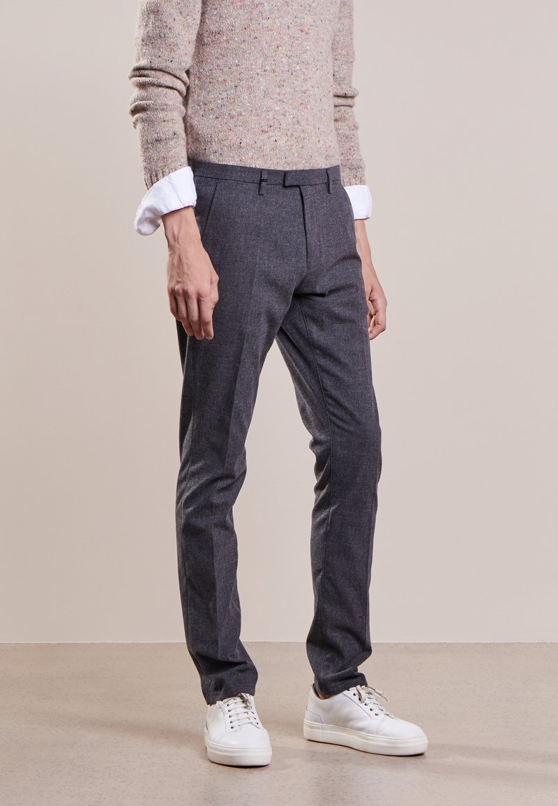 Pantaloni grigi su misura con una texture liscia e pieghe stirate, abbinati a un maglione beige in maglia e scarpe da ginnastica bianche.